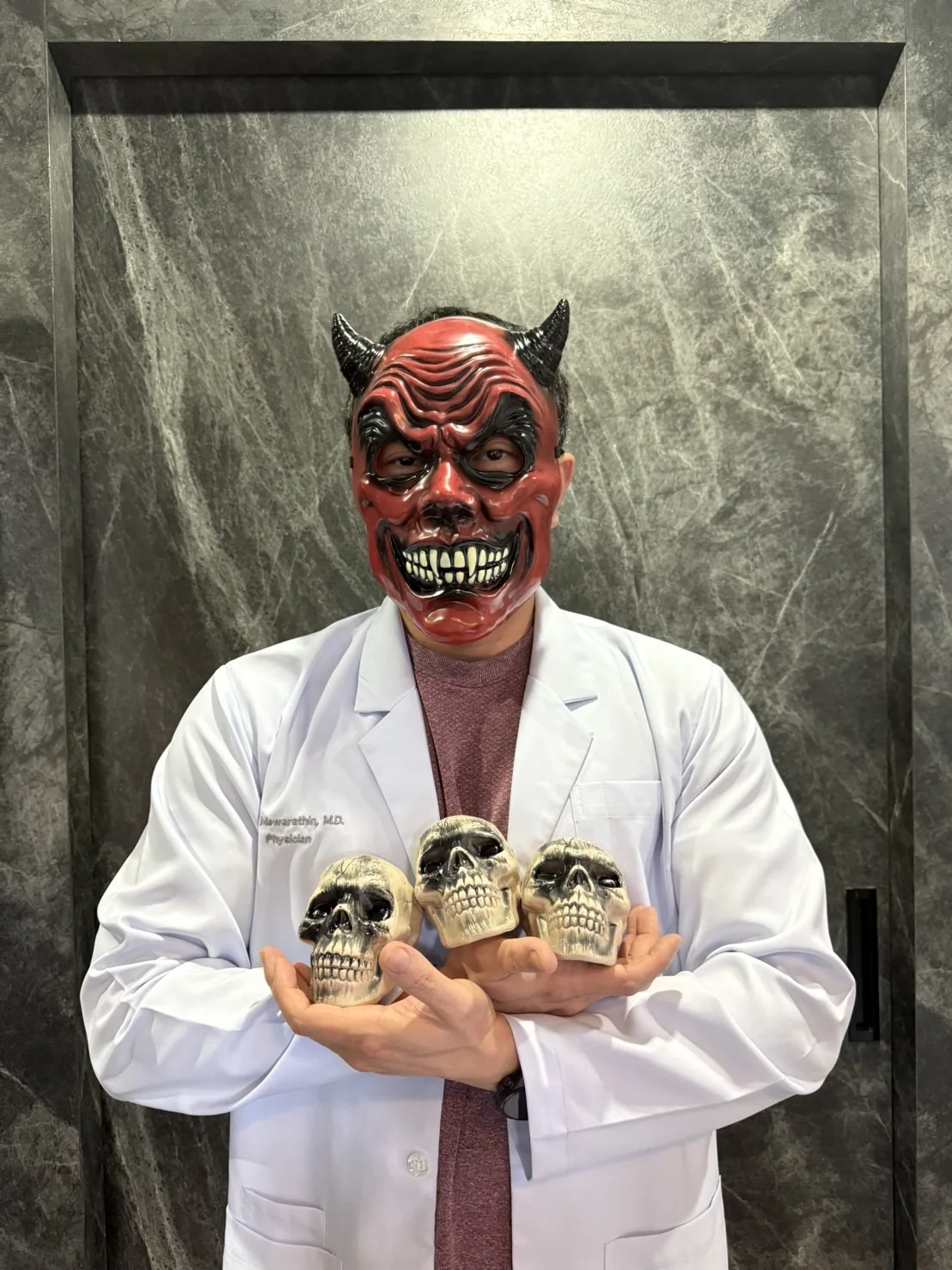 Dr. Krit in a white physician's coat and a full red devil mask with horns, holding three toy skulls against a dark marble wall